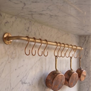 May include: A horizontal brass kitchen rail mounted to a wall, featuring a series of matching brass S-shaped hooks. Three copper-toned metal pans with long handles are suspended from the hooks, hanging vertically against the wall surface.