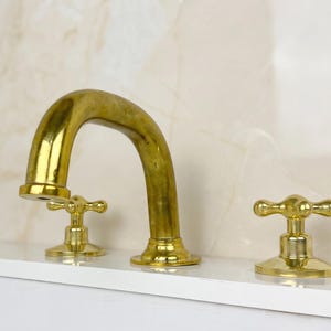 May include: A gold-colored bathroom faucet with a curved spout and two separate handles. The faucet is made of a shiny, metallic material and is mounted on a white surface. The background features a light-colored, textured wall.