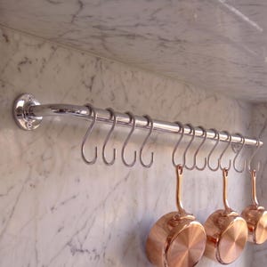 May include: A polished chrome kitchen rail mounted horizontally with multiple silver S-shaped hooks. Three small, round copper pans with long handles are suspended from the hooks, showcasing a metallic finish and a circular base design.