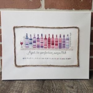 Op de afbeelding: Een rechthoekig wit canvas met een kruissteekborduurwerk omlijst door touw. Het ontwerp toont een wijnglas, twaalf flessen met patronen in roze, paarse en blauwe tinten, de tekst "Deborah", "ST THOMASSON" en "Aged to perfection since 1968".
