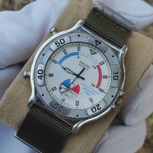 Op de afbeelding: Een Seiko Quartz Yacht Timer Sports 150 horloge met een witte wijzerplaat, blauwe, rode en zwarte accenten. Het horloge heeft een zilveren lunette en een groene nylon band. De wijzerplaat toont de tekst "SEIKO QUARTZ".