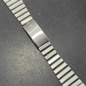 May include: A silver-coloured watch strap with a brushed metal finish. The strap features a series of rectangular links connected by a folding clasp. The clasp has a small, circular emblem. The strap is designed to attach to a watch.