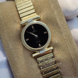May include: A gold-toned Omega De Ville wristwatch with a black dial. The watch features gold hands and hour markers, and a gold expansion band. The watch face displays the Omega logo and "De Ville" text. The watch is on a tan surface.