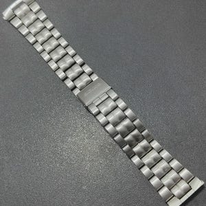 May include: A silver-toned watch band with a brushed metal finish. The band is composed of rectangular links connected by smaller, square-shaped links. A clasp with engraved text is visible. The band is designed to be adjustable and is suitable for various watch sizes.