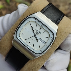 Puede incluir: Un reloj automático Seiko vintage con esfera blanca, manecillas negras y visualización de día y fecha. El reloj tiene una caja rectangular plateada y una correa de cuero negro. La esfera muestra la hora en formatos de 12 y 24 horas.