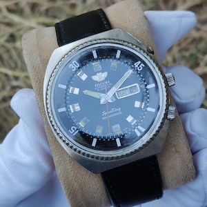 Op de afbeelding: Een vintage Ricoh Space King horloge met een zwarte wijzerplaat, zilveren lunette en zwarte leren band. De wijzerplaat toont de dag en datum, met de tekst "Ricoh Automatic 21 Jewels" en "Space King 100% Water Proof".