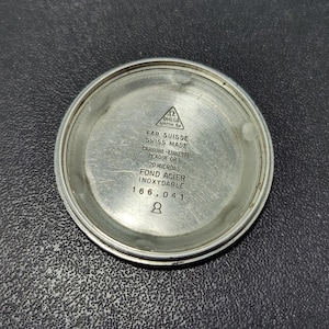 May include: A silver-toned watch back with engraved text, including "OMEGA WATCH Co" and "FAB. SUISSE SWISS MADE." The text also indicates "20 MICRONS" and "INOXYDABLE." The watch back has a circular shape.