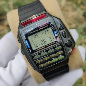 Op de afbeelding: Een zwarte Casio digitale horloge met rekenmachine- en afstandsbedieningsfuncties. Het horloge toont de tijd, dag en datum op een digitaal scherm. Het heeft een zwarte band en knoppen zoals "TV/VOL" en "POWER". De horloge is ongeveer 4 cm breed.