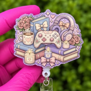 May include: A purple and pink badge reel featuring a stack of books, a video game controller, headphones, a coffee mug, and candles. The design includes floral and bow accents, with a glittery border and a clear plastic reel.