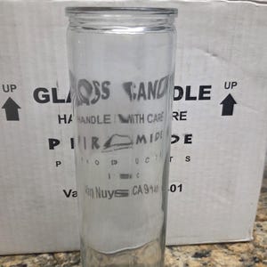 May include: A tall, clear, cylindrical glass candle holder with a smooth surface and a thick base. The glass is transparent, allowing a clear view through the vessel. It features a simple, functional design intended for holding a standard pillar candle.