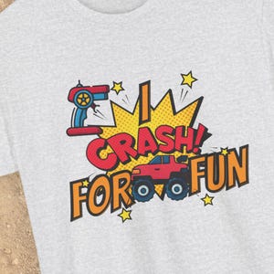 Peut inclure: T-shirt gris clair avec un graphisme coloré. Le motif présente une télécommande rouge et bleue, un monster truck rouge et les mots "I CRASH! FOR FUN" en gras, dans un style bande dessinée. Des étoiles jaunes et une explosion jaune complètent le design.