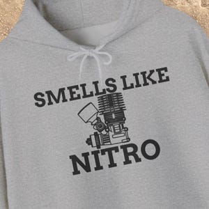 May include: A light gray hoodie with the text "SMELLS LIKE NITRO" in black, along with a graphic of an engine. The hoodie has a drawstring and a front pocket, perfect for casual wear.