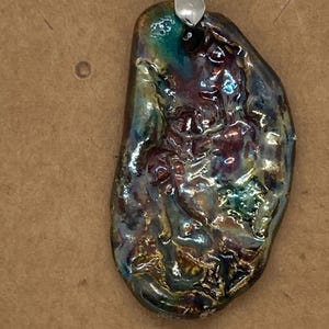 May include: A handcrafted ceramic pendant with a unique, organic shape. The pendant features a glossy, iridescent glaze with shades of teal, purple, and gold. It is suspended from a silver-toned bail and a white beaded chain, creating a one-of-a-kind jewelry piece.