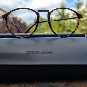 May include: Round eyeglasses with a brown and black tortoiseshell pattern frame resting on a black hard-shell case. The case features the embossed text GIORGIO ARMANI on the front center. The glasses have thin metal temples and a bridge.