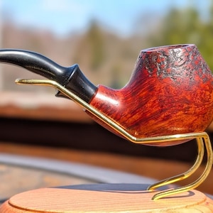 May include: A handcrafted tobacco pipe with a dark, curved stem and a reddish-brown bowl. The bowl is supported by a gold-colored metal stand, and the pipe rests on a wooden base. The pipe's bowl has a textured surface.
