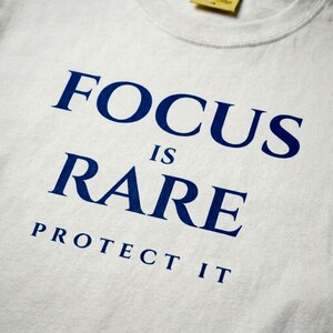 K&ouml;nnte beinhalten: Wei&szlig;es T-Shirt mit dem marineblauen Text "FOCUS IS RARE PROTECT IT". Der Text ist zentriert und in einer fetten, serifenlosen Schriftart. Das Shirt hat einen Rundhalsausschnitt und ein gelbes Etikett.