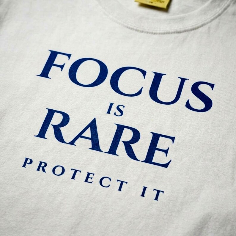 K&ouml;nnte beinhalten: Wei&szlig;es T-Shirt mit dem marineblauen Text "FOCUS IS RARE PROTECT IT". Der Text ist zentriert und in einer fetten, serifenlosen Schriftart. Das Shirt hat einen Rundhalsausschnitt und ein gelbes Etikett.