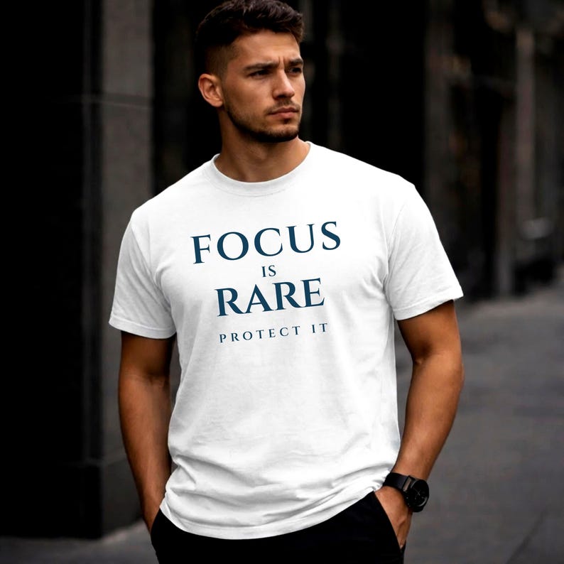 K&ouml;nnte beinhalten: Wei&szlig;es T-Shirt mit dem marineblauen Text "FOCUS IS RARE PROTECT IT". Das Shirt hat kurze &Auml;rmel und scheint aus einem weichen Material zu sein. Die Person tr&auml;gt eine schwarze Uhr.