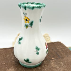 May include: A small, white ceramic vase with a green scalloped rim and base. The vase is decorated with hand-painted yellow flowers, green leaves, and small pink and red floral accents. The vase is approximately 5 inches tall.