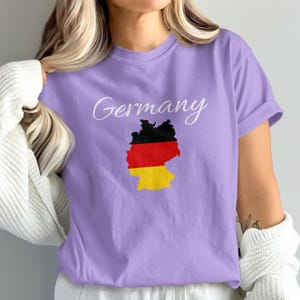 May include: A lilac-colored t-shirt with the word "Germany" in cursive script. Below the text is a graphic of the German flag in the shape of the country. The flag colors are black, red, and yellow.