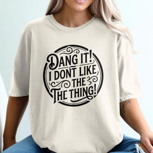 May include: Cream-colored t-shirt with a black circular graphic that reads "Dang It! I Don't Like The Thing!" in a bold, stylized font. The shirt has short sleeves and a crew neck.