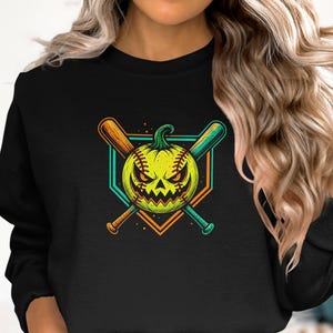 May include: A black crewneck sweatshirt featuring a graphic print of a yellow pumpkin with a baseball-style stitch pattern and a menacing face. The pumpkin is positioned in front of two crossed baseball bats and a geometric diamond-shaped frame.