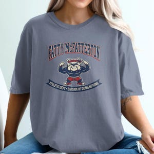 Könnte beinhalten: Ein schieferblaues Kurzarm-T-Shirt mit einer Grafik eines muskulösen Bulldogs, der ein rotes Stirnband und eine rote Hose trägt. Der Text FATTY MCFATTERTON steht darüber und ATHLETIC DEPT • DIVISION OF DOING NOTHING auf einem Banner darunter.