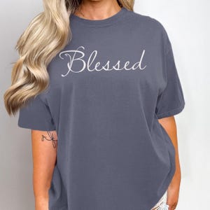 May include: A slate blue t-shirt with the word "Blessed" in a white script font. The short-sleeved shirt has a crew neck and a relaxed fit. The text is centered on the front of the shirt.