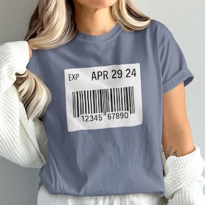 Peut inclure: T-shirt bleu avec un grand code-barres blanc. Le texte indique "EXP APR 29 24" et "12345 67890". Le t-shirt a des manches courtes et une coupe décontractée.