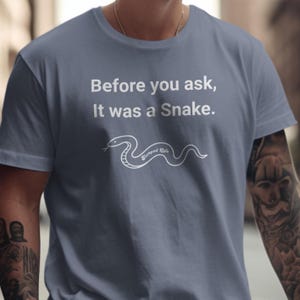 Puede incluir: Una camiseta de manga corta color azul pizarra con el texto impreso en blanco 'Before you ask, It was a Snake.' sobre una ilustración de líneas blancas de una serpiente con las palabras 'Stumped Life' en cursiva sobre su cuerpo.