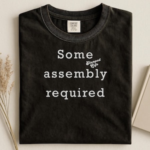 May include: Black t-shirt with the words "Some assembly required" in white, along with the phrase "Slumped Life". The shirt has a crew neck and a tag that says "Comfort Colors".