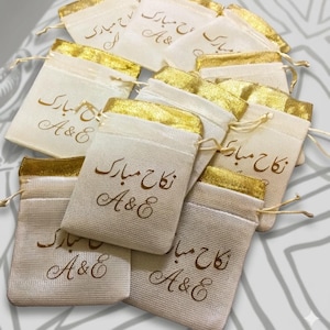 Op de afbeelding: Een verzameling kleine, off-white stoffen buideltjes met een trekkoord en een metallic gouden bovenrand. Elk buideltje is aan de voorzijde bedrukt met de Arabische tekst "نکاح مبارک" en de letters "A&E" in een donkerbruin schrijflettertype.