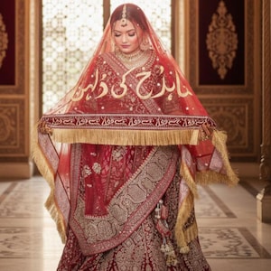 May include: A woman in a traditional red wedding dress with gold embroidery and a matching veil. The veil has gold text and a gold fringe. The dress features intricate patterns and a long skirt, creating an elegant and cultural look.