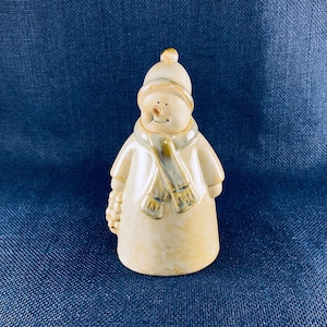 May include: A ceramic snowman figurine with a cream-colored body, wearing a matching hat and coat. The snowman has a scarf with a blue and yellow pattern, a carrot nose, and a small basket on one arm. The figurine is a festive winter decoration.