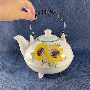 May include: White ceramic teapot with a black and metal handle, featuring a hand-painted design of two yellow sunflowers with brown centers and green leaves. The teapot has a white lid with a green trim and three small feet.