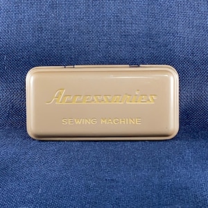 May include: A tan metal sewing machine accessories box. The box has rounded edges and a hinged lid. The word "Accessories" is written in a cursive font, and "SEWING MACHINE" is written in a sans-serif font.