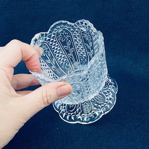 May include: Clear glass dish with a scalloped rim and floral and diamond pattern. The dish sits on a matching base with a similar design. The glass is held by a hand against a dark blue background.