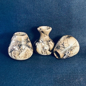 May include: Three small, beige ceramic vases with black lightning-like patterns and white geometric designs. The vases have varying shapes, including a bulbous form and a narrow-necked vase. The background is a dark blue.