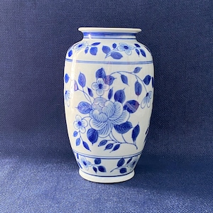 May include: A white ceramic vase with blue floral patterns. The vase features a large blue flower and smaller blue flowers with leaves. The top and base of the vase have blue accents. The vase is on a blue surface.