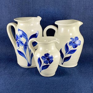 May include: Three off-white ceramic pitchers with blue floral designs. The pitchers vary in size, with a large one on the left and a smaller one in the center. The blue flowers and leaves are hand-painted. The background is a textured blue fabric.