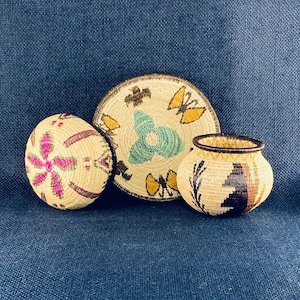 May include: Three woven baskets with detailed patterns. The largest is a round, flat basket with butterfly and floral designs in yellow, green, and brown. A smaller round basket features a pink floral design. The smallest is a round pot with black and brown patterns.