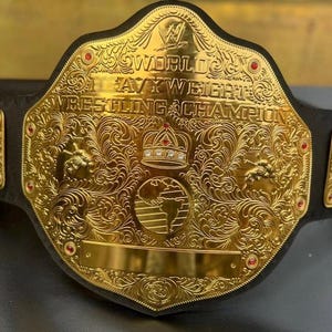 以下が含まれることがあります： 金色のレスリングチャンピオンシップベルト。複雑な花柄とスクロールワークのデザインが施されています。ベルトには「World Heavyweight Wrestling Champion」の文字と王冠が描かれています。赤い宝石が縁にセットされています。ベルトは黒い革のストラップに付いています。
