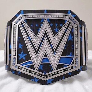 May include: A professional wrestling championship belt with a black and blue star pattern. The belt features a large silver "W" logo encrusted with rhinestones, and the words "WORLD HEAVYWEIGHT CHAMPION" are displayed.
