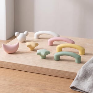 Pode incluir: Uma coleção de ferragens para móveis incluindo um puxador branco em forma de rato, um puxador rosa em forma de meia-lua, dois puxadores em forma de estrela em amarelo e verde, e quatro puxadores arqueados em branco, rosa, amarelo e verde mate.