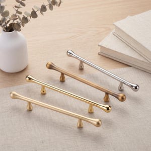 May include: Four elongated cabinet pulls arranged in a row, featuring a smooth, rounded bar design with two small, teardrop-shaped mounting posts. The set includes one polished silver finish, one brushed bronze, one polished gold, and one matte gold.