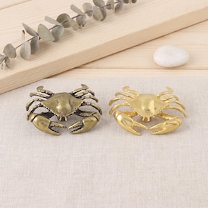 May include: Two crab-shaped cabinet knobs. One knob features an antique brass finish with dark, weathered detailing, while the other knob has a bright, polished gold finish. Both knobs display detailed legs and large, prominent front claws.