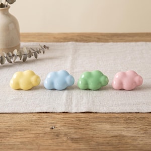 May include: Four small, smooth, cloud-shaped ceramic objects arranged in a horizontal row. The colors from left to right are pastel yellow, light blue, soft green, and pale pink. Each piece has a glossy finish and a rounded, puffy appearance.