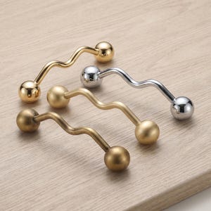 May include: Four wavy cabinet handles with spherical ends, arranged in a diagonal row. The set includes one polished gold handle, one polished silver handle, and two matte brass handles, each featuring a distinct undulating, serpentine shape.