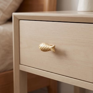 May include: A light-toned wooden nightstand featuring a single drawer with a gold-colored metal cabinet knob shaped like a fish. The fish knob has a textured scale pattern on its surface and is centered on the front panel of the drawer.