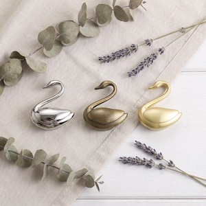 May include: Three swan-shaped decorative objects arranged in a row. The first swan is polished silver, the second is a brushed antique bronze, and the third is a matte gold finish. Each swan features a curved neck and a smooth, rounded body.